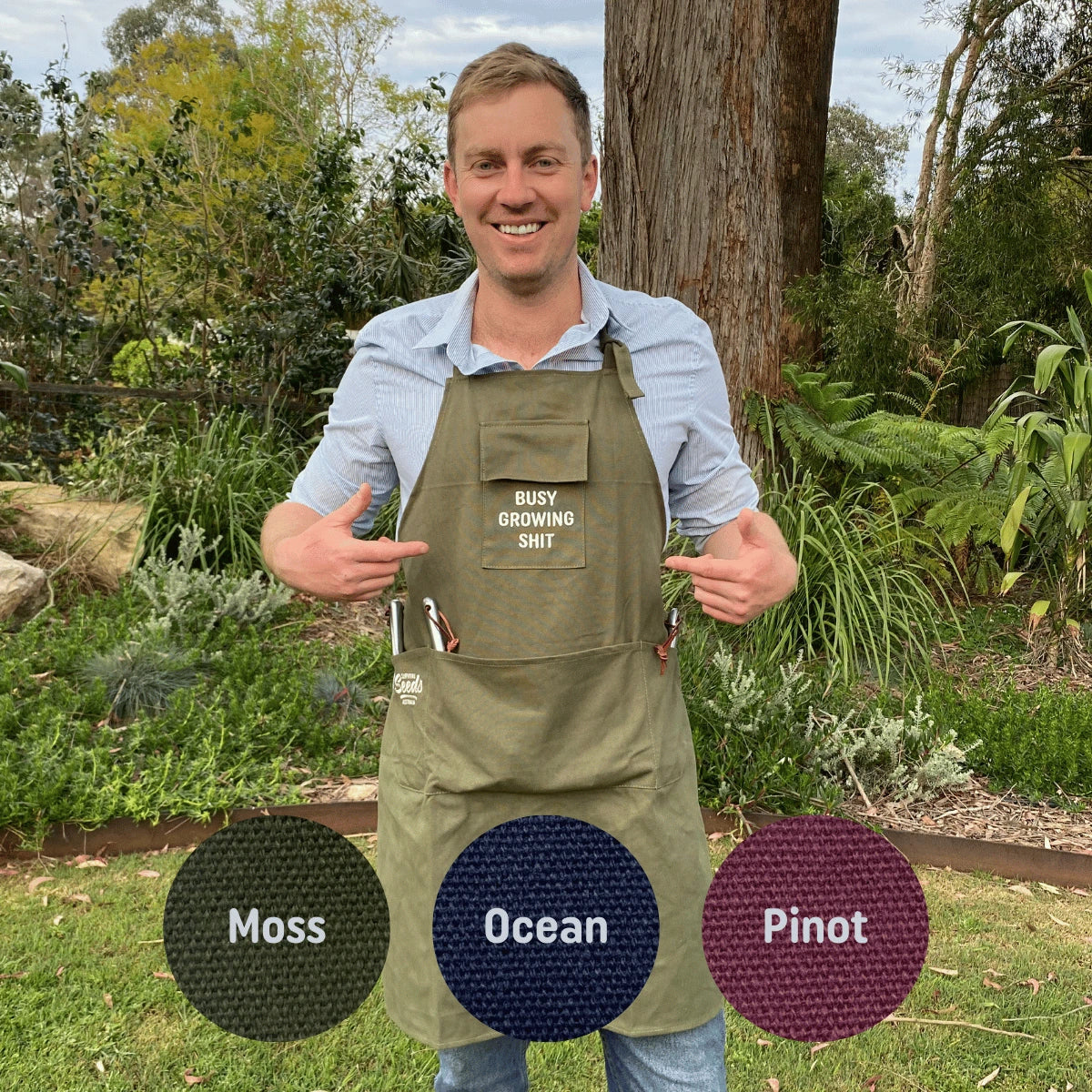 Man wearing a green apron with text, standing outdoors in a garden setting.