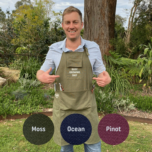 Man wearing a green apron with text, standing outdoors in a garden setting.